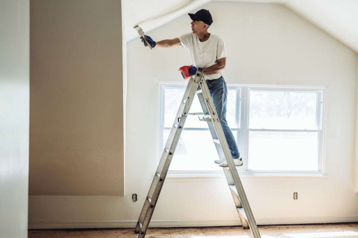 Persona de pie en una escalera pintando la parte superior de una pared interior con un rodillo de pintura
